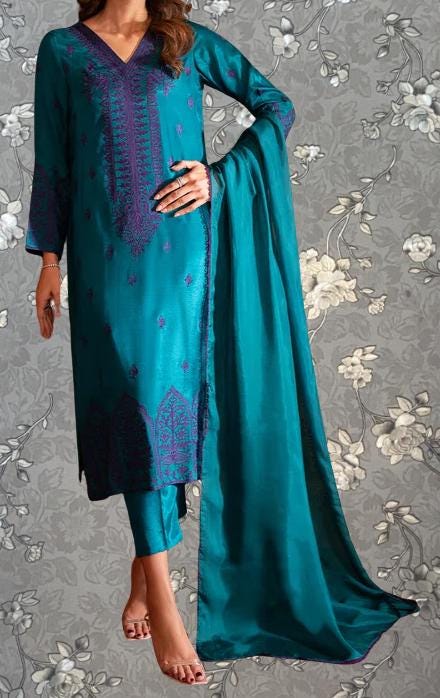 A woman wearing a teal and purple dress with intricate embroidery, standing against a gray floral background.