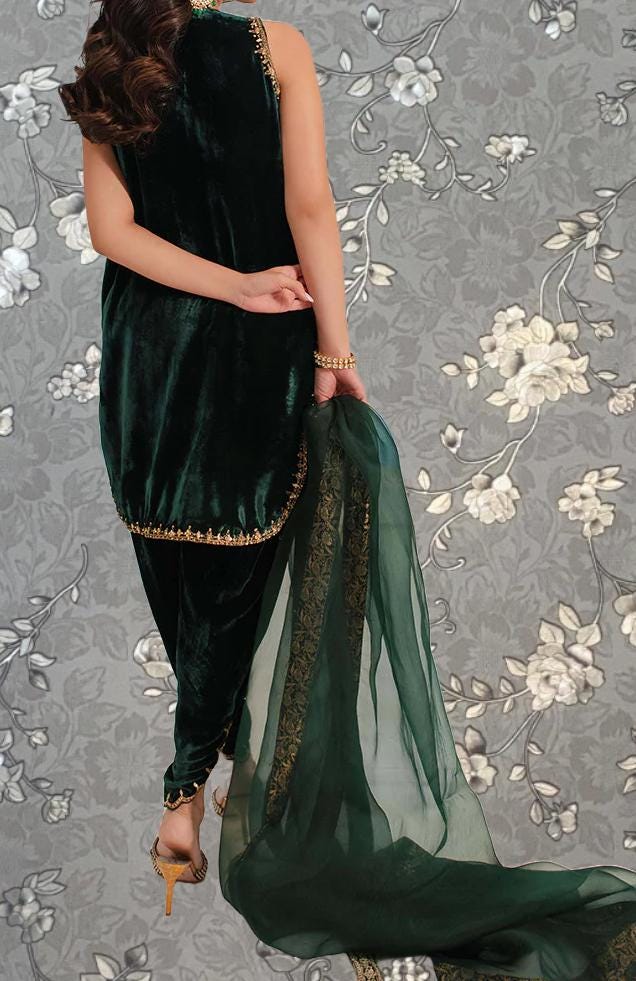 A woman wearing a green velvet dress with gold trim and a matching green shawl is standing against a gray floral background.