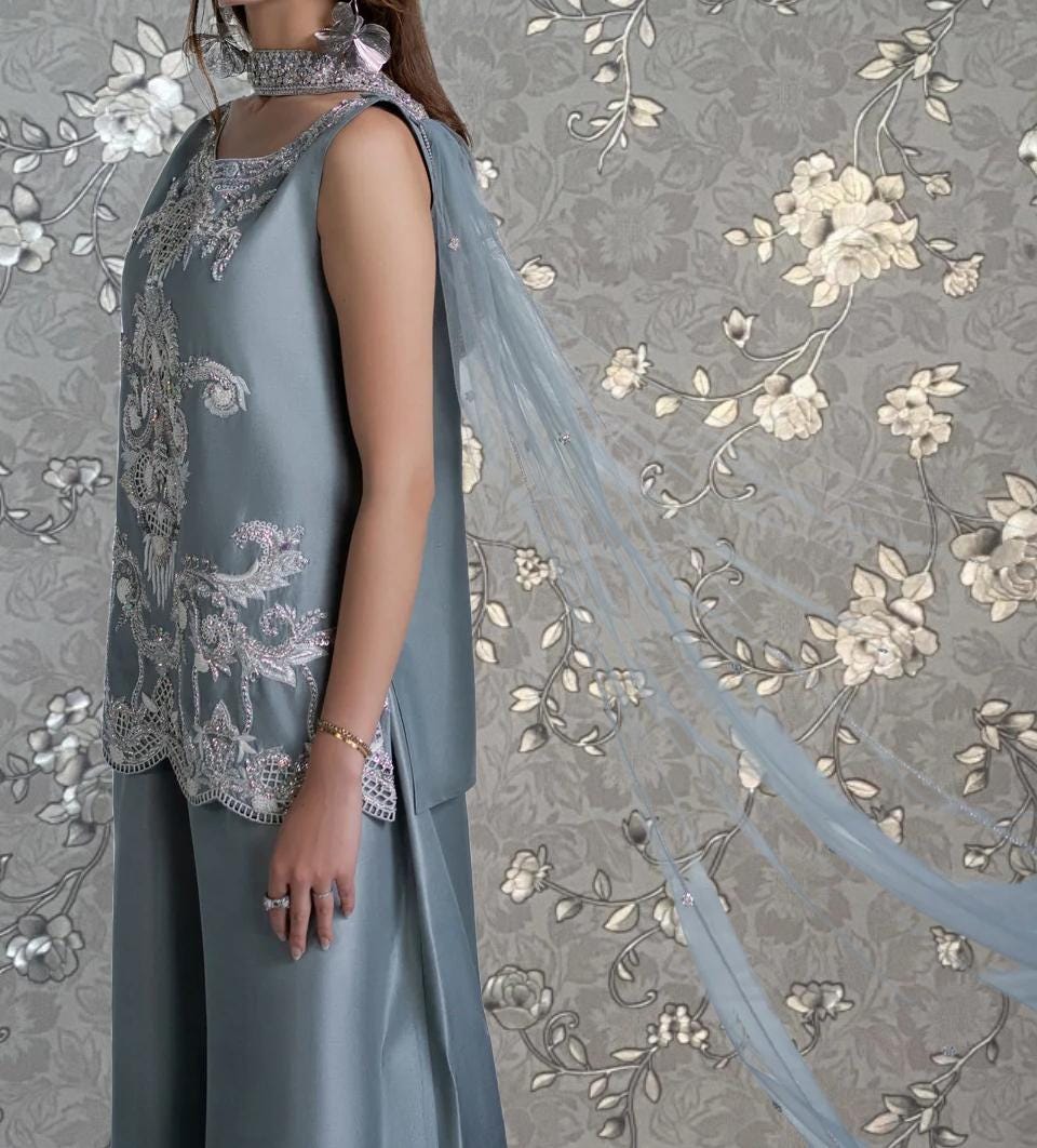 A woman wearing a gray dress with intricate embroidery stands in front of a floral patterned background.