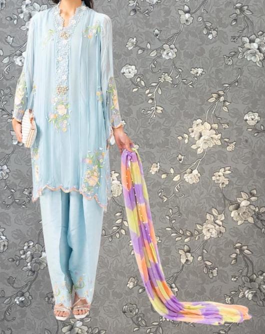 A woman wearing a light blue dress with floral embroidery and holding a colorful scarf stands against a gray floral background.