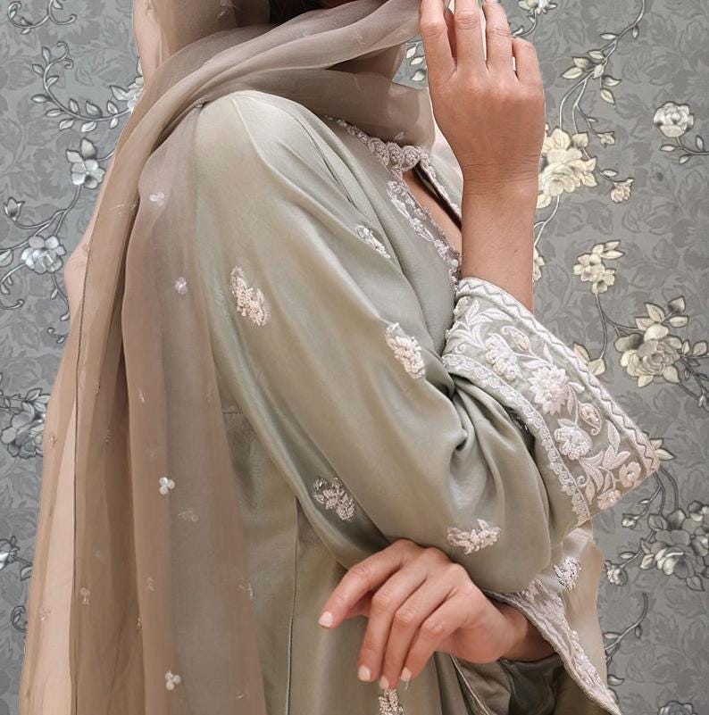 A woman wearing a light green dress with intricate embroidery stands against a floral patterned background.
