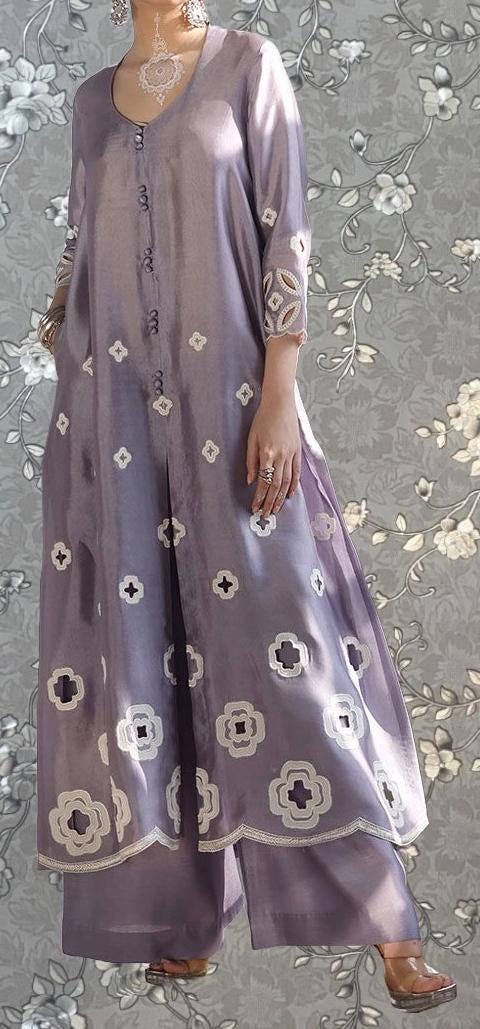 A woman wearing a long purple dress with white floral embroidery stands against a gray floral background.