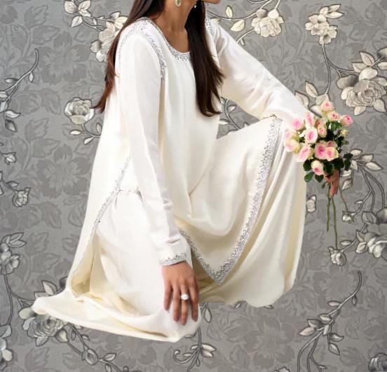 A woman in a white dress holding a bouquet of pink flowers stands against a gray floral background.