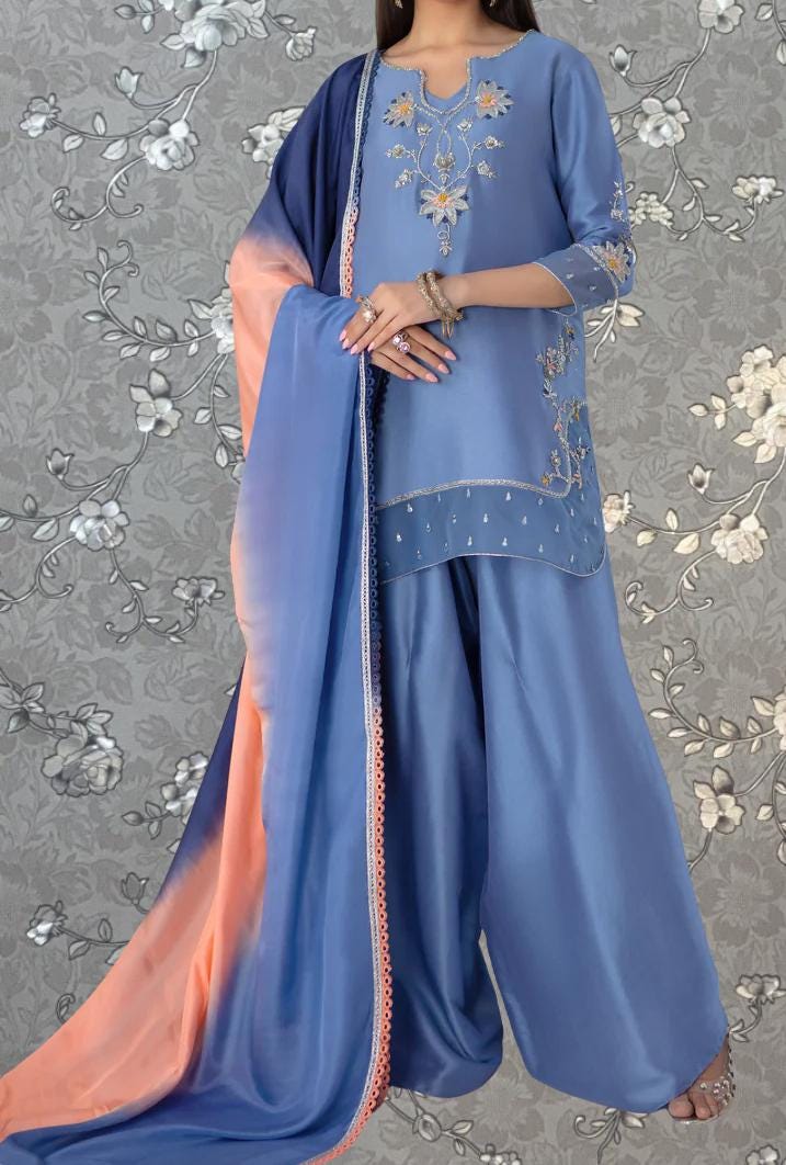 A woman wearing a blue and orange dress with intricate embroidery, standing against a gray floral background.