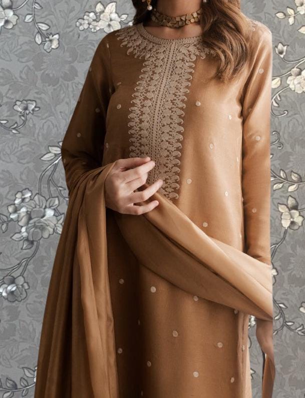 A woman wearing a brown dress with a floral pattern and a matching shawl stands against a gray floral background.