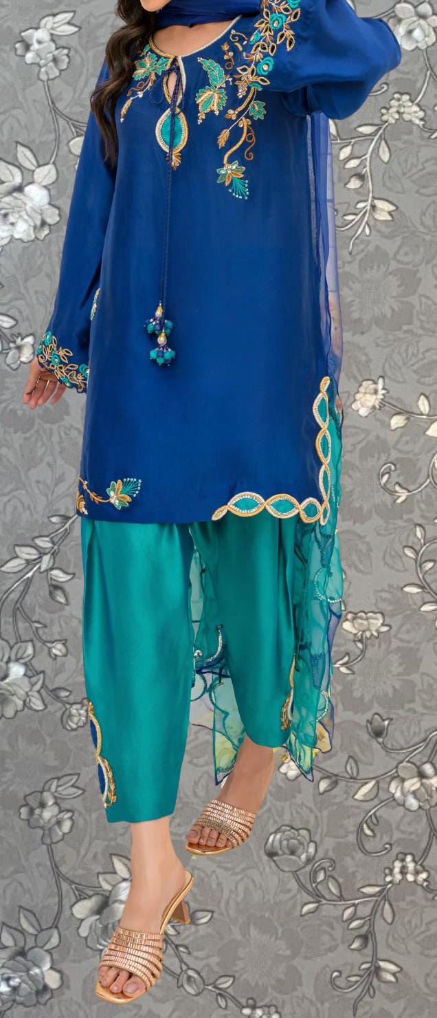 A woman wearing a blue dress with intricate embroidery and a matching blue skirt, standing against a gray floral background.