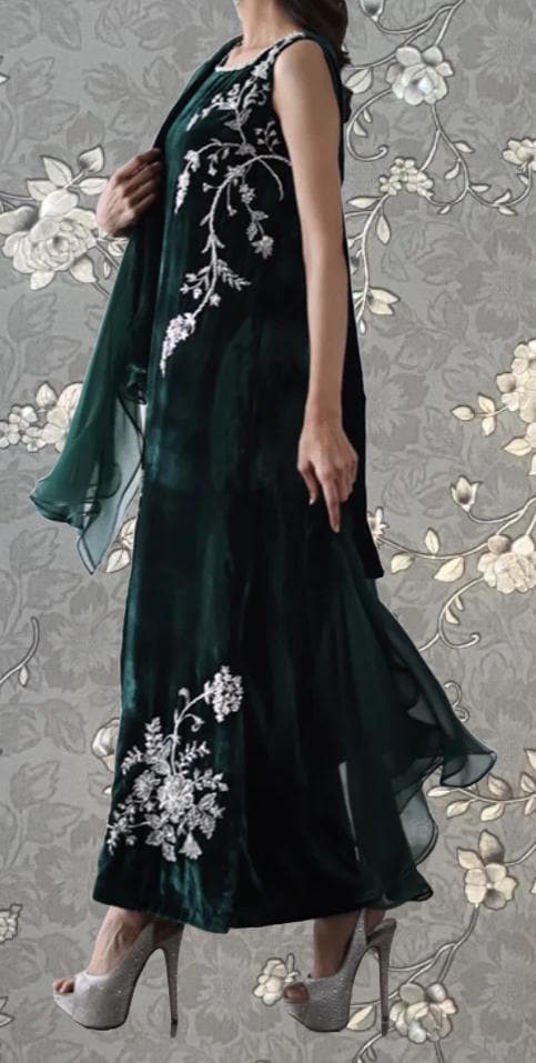 A woman wearing a long, green dress with white floral embroidery stands against a floral background.