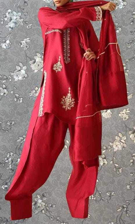 A person wearing a red outfit with intricate embroidery stands against a gray floral background.