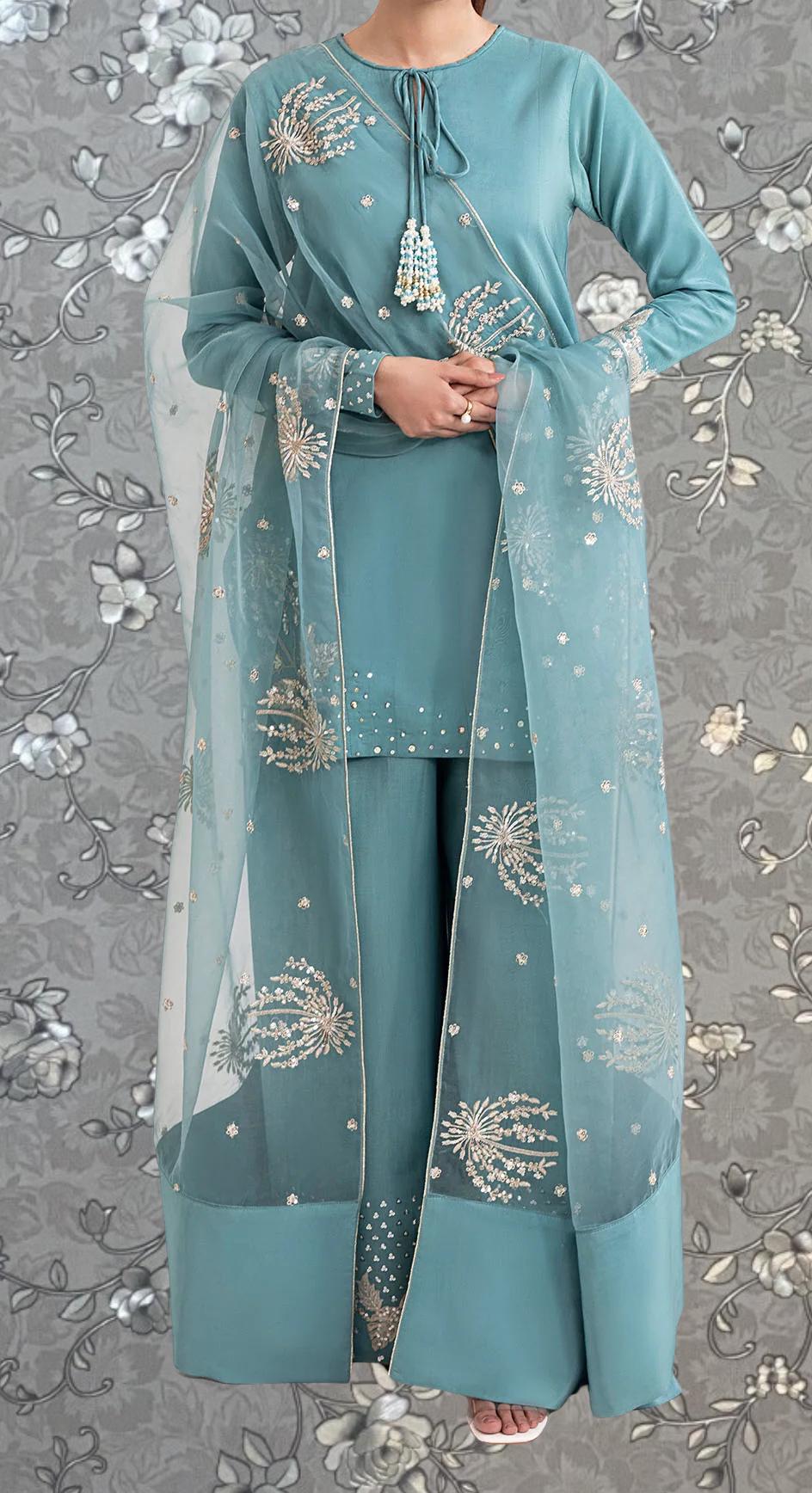 A woman wearing a teal blue dress with intricate embroidered designs, including white flowers, stands against a gray floral background.