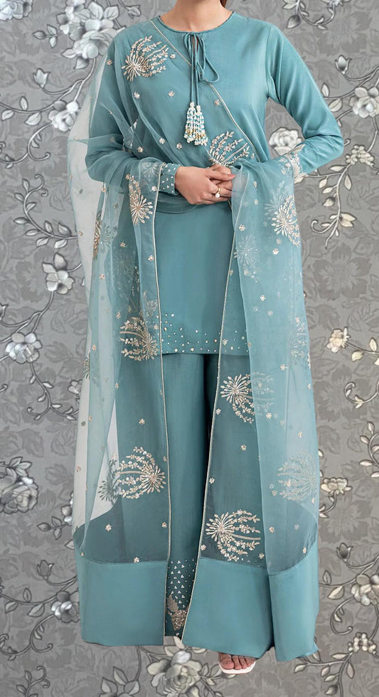 A woman wearing a teal blue dress with intricate embroidered designs, including white flowers, stands against a gray floral background.