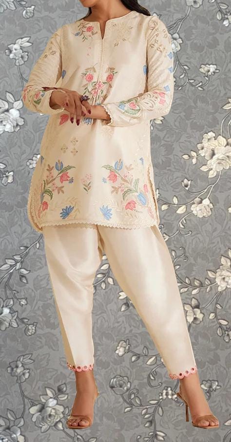 A woman wearing a cream-colored dress with floral embroidery stands against a gray floral background.