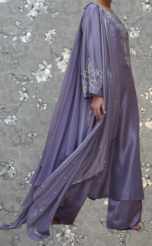 A person wearing a long, flowing purple dress with intricate embroidery stands against a floral background.