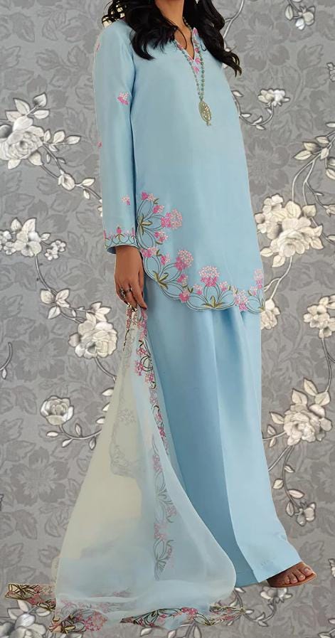 A woman wearing a light blue dress with floral embroidery, standing against a gray floral background.