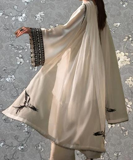 A person wearing a long, flowing white robe with black accents, including a hood and a bird design, stands against a gray floral background.