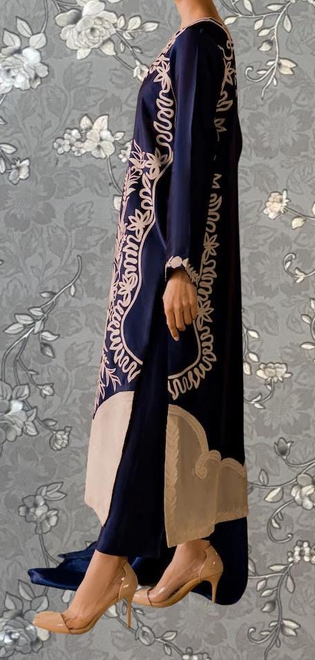 A person wearing a long, navy blue dress with intricate white embroidery stands against a gray floral background.