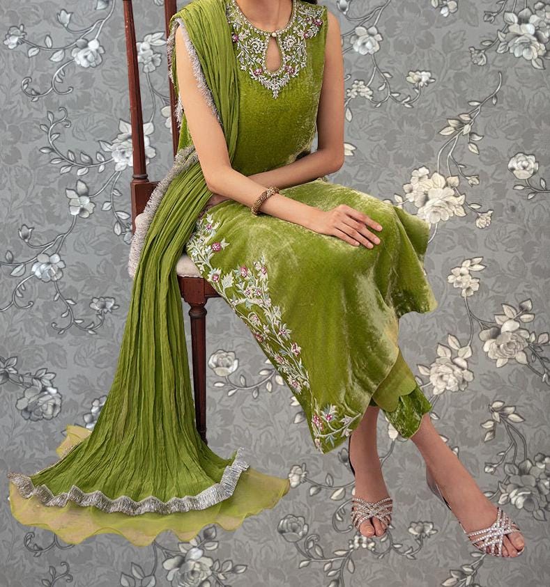 A woman wearing a green dress with floral embroidery is sitting on a chair against a gray floral background.